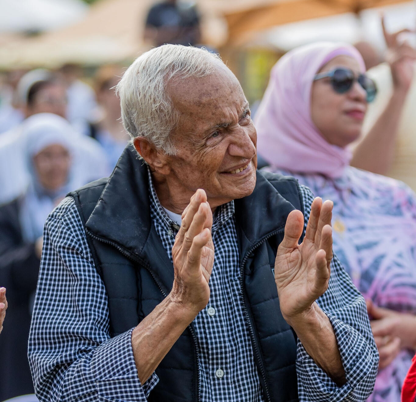 An older man with white hair is standing and smiling while clapping. In the background, several people of different ages are also standing, all looking in the same direction.