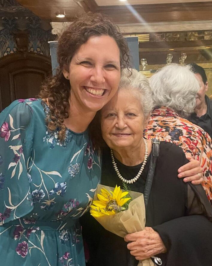 Dina Hashish in a teal floral dress standing beside an older woman with short white hair, who is holding a small bouquet of sunflowers.